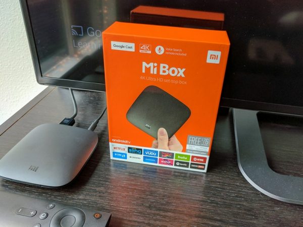 Подключённая к телевизору приставка Xiaomi Mi TV Box лежит на тумбе рядом с телевизором и упаковочной коробкой